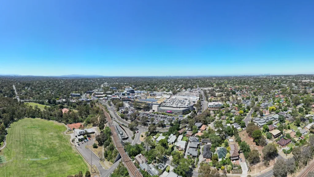 An aerial view over Greensborough in Melbourne's north-east, photographed on 12 December 2024, showing the Greensborough Plaza and its car parks, the railway line and station, a sports oval, tree-lined streets and suburban rooftops.