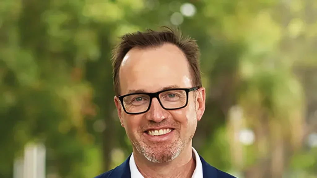 David Shoebridge in a navy blazer and glasses, smiling, photographed outdoors against green trees in 2025.