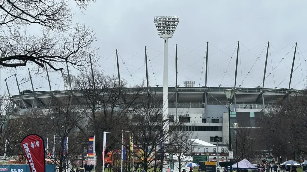 Crowds walking past bare winter trees towards the Melbourne Cricket Ground and its floodlight tower before the 2026 AFL Grand Final.