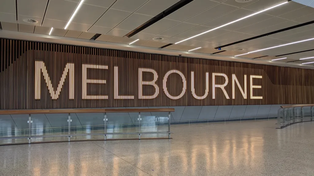 An empty international arrivals hall at Melbourne Airport, with the illuminated word MELBOURNE set into a timber slat wall above a glass balustrade.