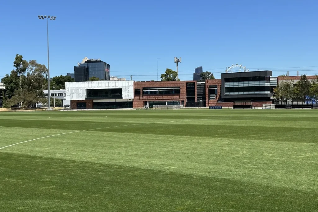 Arden Street Oval in North Melbourne on a clear day, with the empty mown ground in the foreground, the club's red-brick headquarters across the far side and city towers behind.