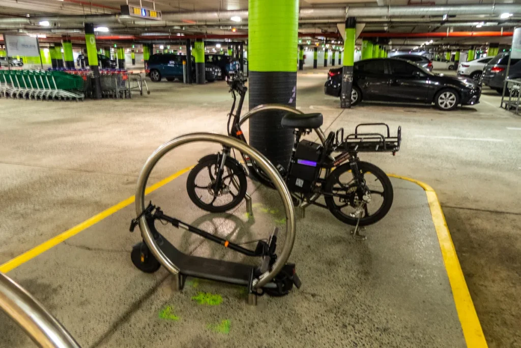 A folded e-scooter and an e-bike with a rear rack, locked to a stainless-steel hoop bike rail in an underground shopping-centre car park in Chermside, Brisbane. Illustrative; not connected to the seizures.