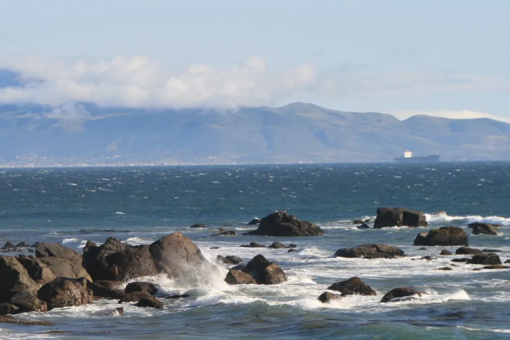 Waves breaking over dark rocks on the Baja California coast near Ensenada, with a ship on the horizon and cloud-capped mountains across the bay.