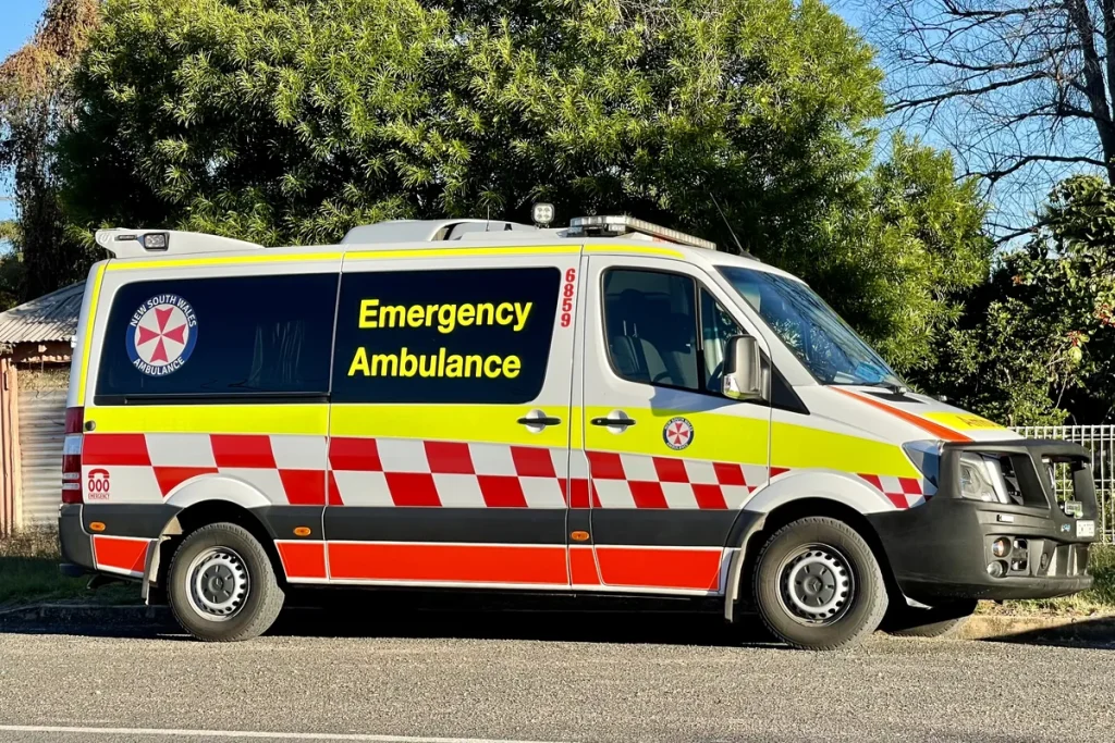 A NSW Ambulance van parked beside a quiet suburban street in late-afternoon sun, with trees, a tin shed and a mesh fence behind it. Illustrative; not taken at Wallsend.