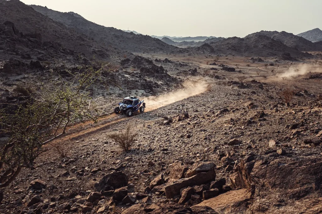 A blue off-road buggy trailing a plume of dust along a stony track through a dry, hilly desert valley at dusk.