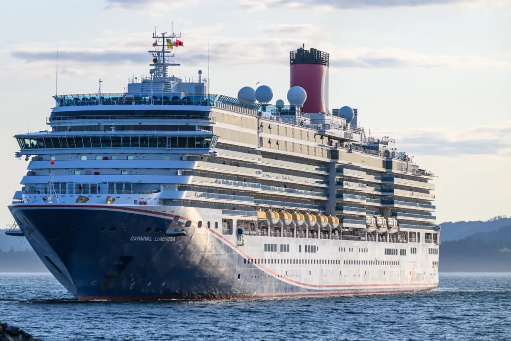 The cruise ship Carnival Luminosa, with a dark blue hull and red funnel, moving through calm water near Victoria, British Columbia, with forested hills behind under a pale evening sky.