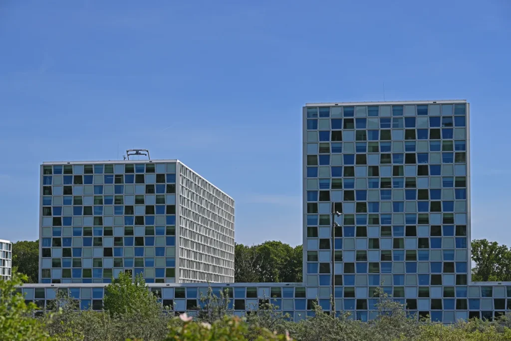 Two tall office towers with chequered blue and grey glass facades against a clear sky, trees and shrubs in the foreground.