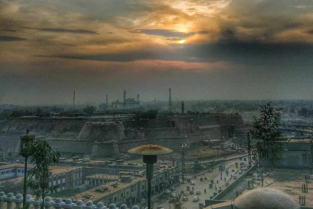 Hazy evening sky over Peshawar, with the walls of the Bala Hisar fort, flat rooftops and a busy street below, seen from a terrace with a white balustrade and lamps.
