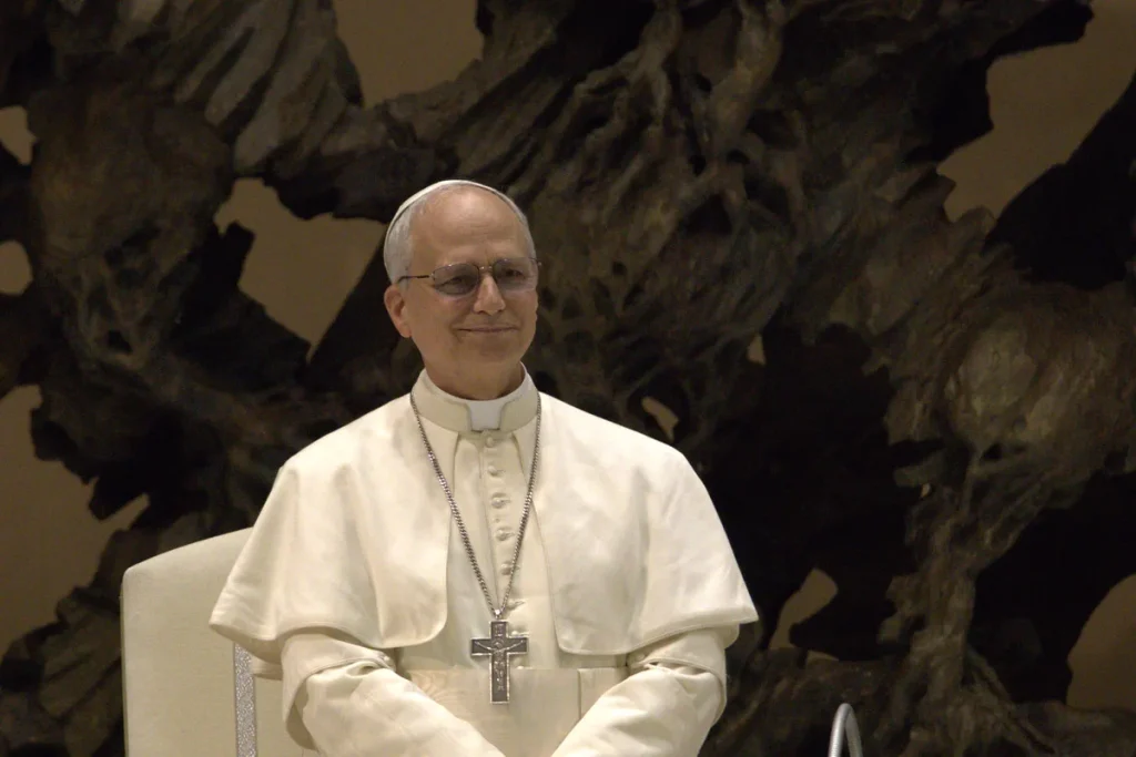 Pope Leo XIV in white cassock and pectoral cross, seated and smiling in front of a large dark bronze sculpture.
