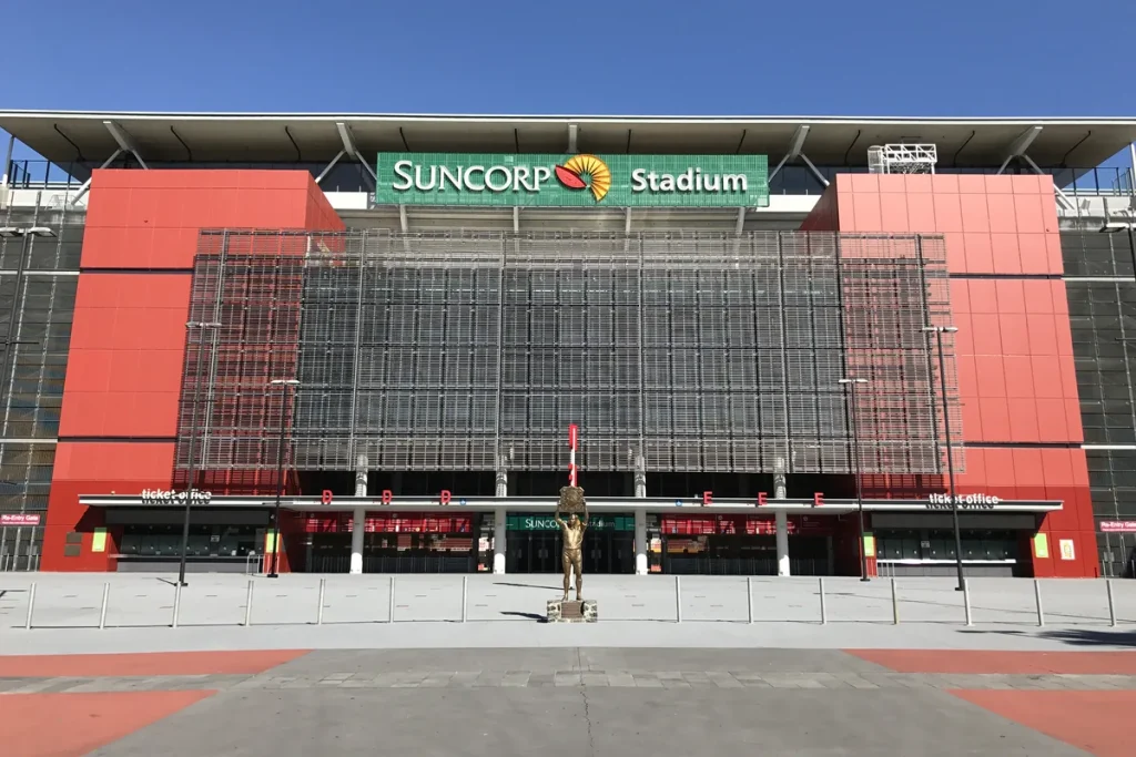 The red and grey Milton Road facade of Suncorp Stadium in Brisbane under a clear blue sky, with the stadium's name sign above an empty forecourt and a bronze statue in front of the ticket office.