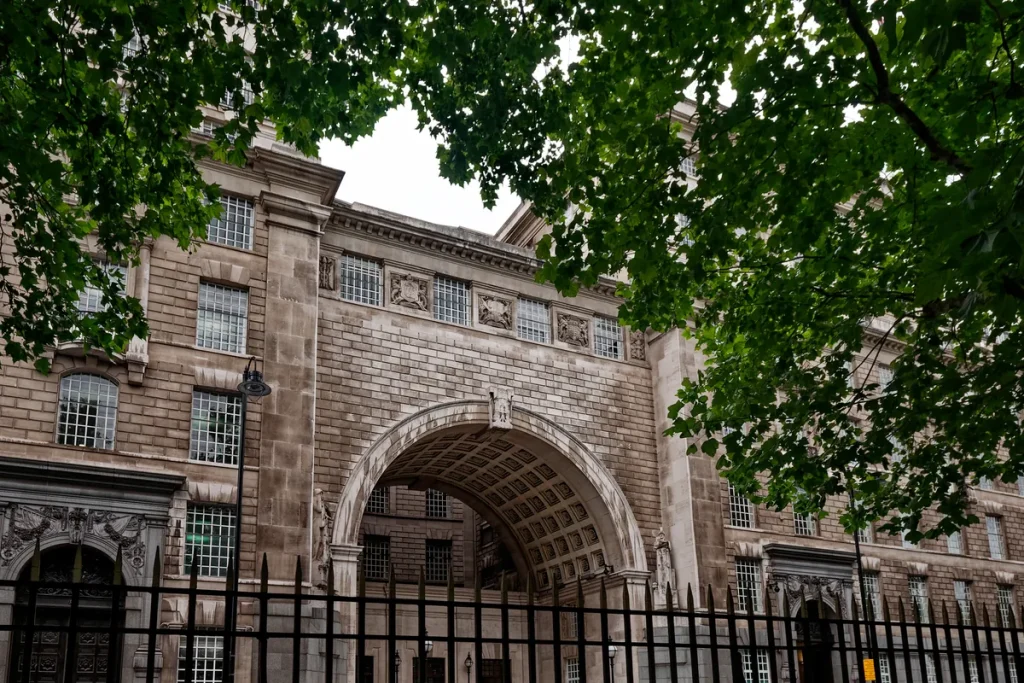 The stone facade and central archway of Thames House on Millbank, London, seen through black iron railings beneath overhanging tree branches.