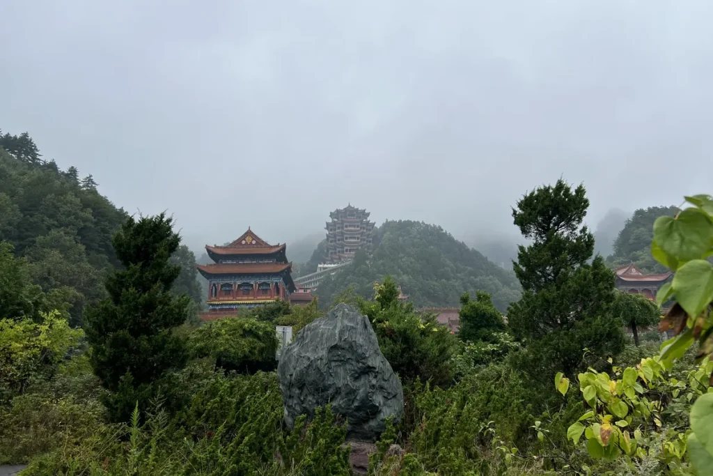 Red and gold temple halls and a tall multi-tiered pagoda on a misty forested hillside, with a large dark rock and green shrubs in the foreground.