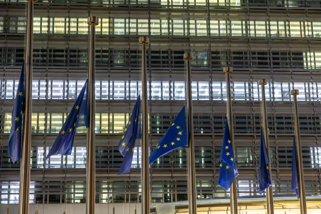 A row of blue European Union flags hanging at half-mast on steel poles in front of a lit glass office facade at dusk.