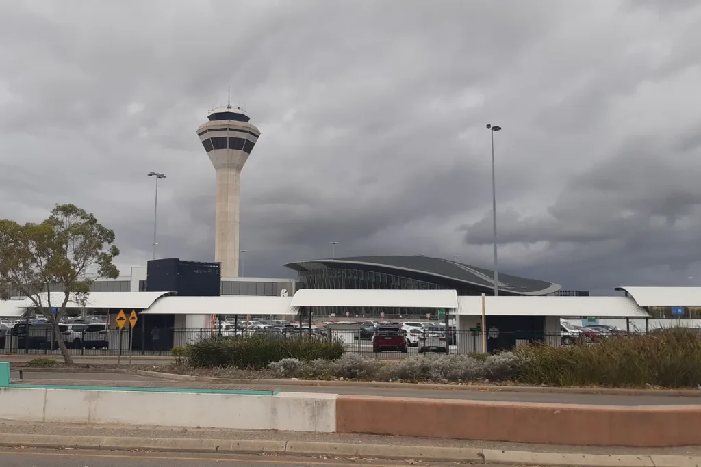 An airport control tower and a curved station roof beyond a car park and covered walkway under heavy grey cloud.