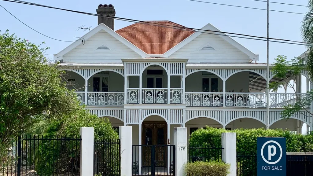 A white two-storey Queenslander house with a red roof and lace-trimmed verandahs behind a front fence, with a for sale sign at the front of the garden.