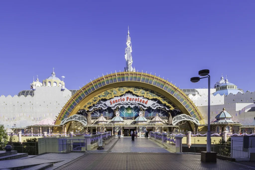 A rainbow-arched theme park entrance with a white spire, pastel domes and a red Sanrio Puroland sign under a clear blue sky.