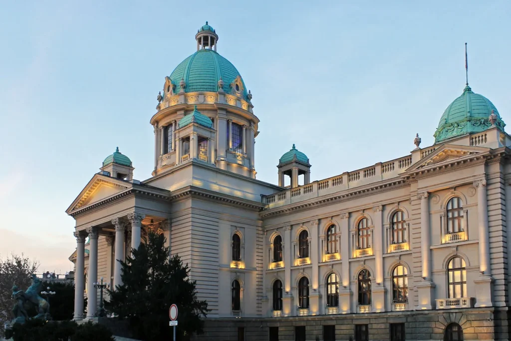 A domed neoclassical parliament building with green copper cupolas and lit arched windows under a pale evening sky.