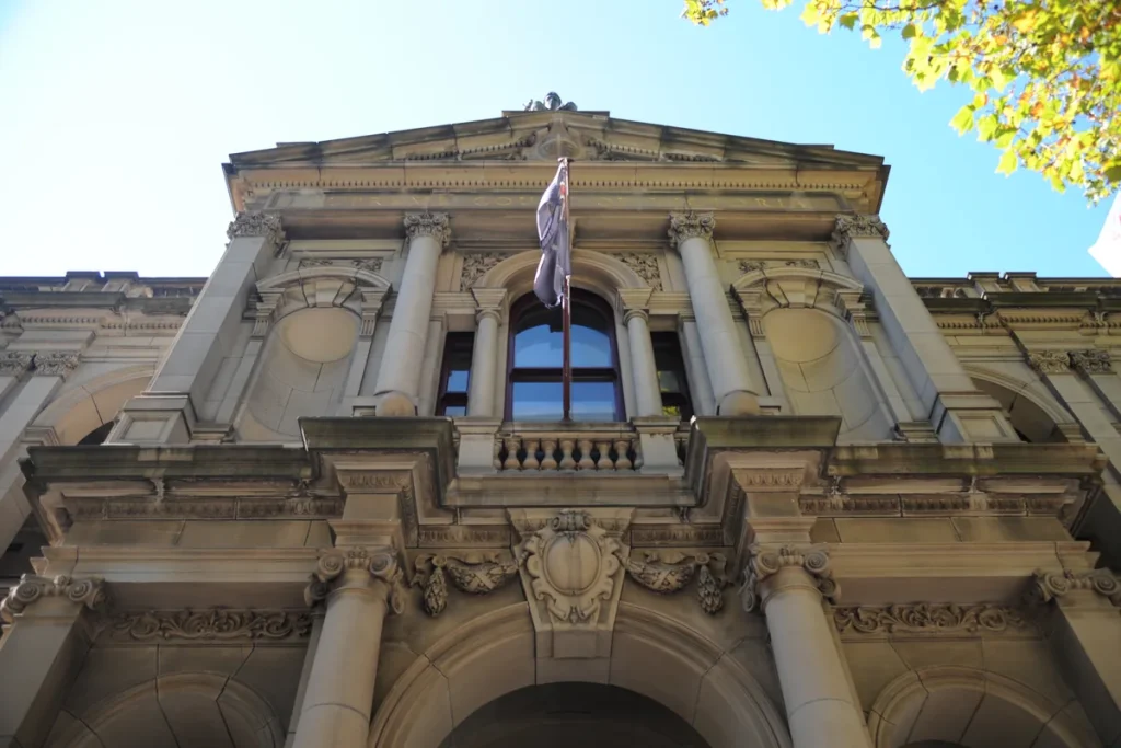 A sandstone court facade seen from below, with carved columns, an arched window and a flag above the entrance against blue sky.