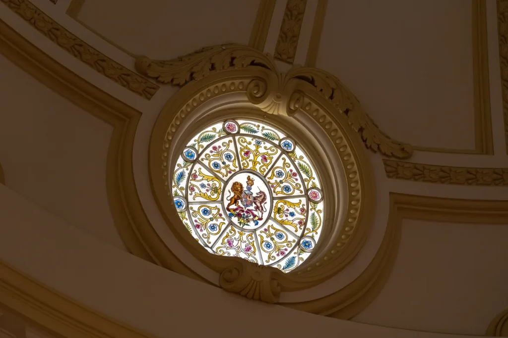 A round stained-glass window bearing a royal coat of arms, set in a cream plaster dome with carved scrolls.