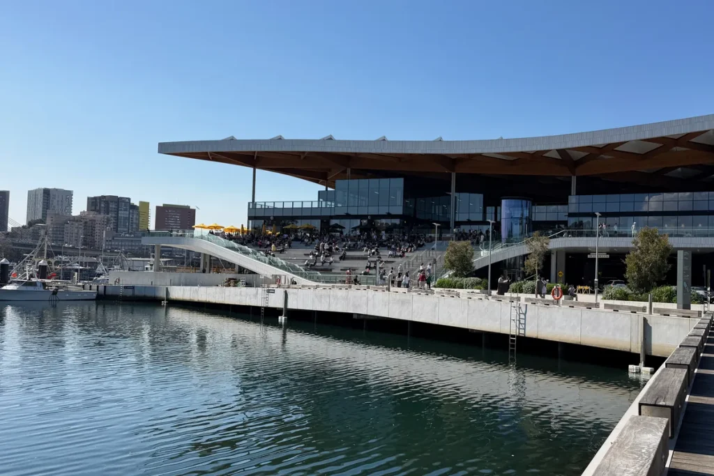 A long low building with a wide timber-ribbed roof on the edge of a calm harbour, city towers behind and a boardwalk at right.